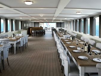 Geräumige Lounge auf der MS Magnifique III mit langen Esstischen, weißen Stühlen und Panoramafenstern auf beiden Seiten. Barbereich im Hintergrund.