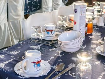 Frühstückstisch im Salon der MS Panagiota mit blauer nautischer Tischdecke, weißem Geschirr, Tassen, Saftgläsern und Getränken.