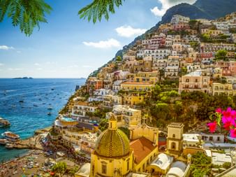 Positano Küstenstadt mit bunten Gebäuden an steilem Hang. Kirche mit gelber Kuppel im Vordergrund, Mittelmeer mit Booten sichtbar.
