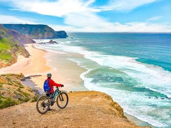 Radfahrer in roter Jacke auf Klippe mit Blick auf Sandstrand und türkisfarbenen Atlantik bei Sagres. Dramatische Küstenklippen rahmen die Bucht.