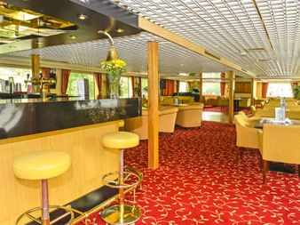 Helle Bar auf der MS Normandie mit Holztheke, gelben Barhockern, gelben Sesseln, rot gemustertem Teppich und weißer Gitterdecke.