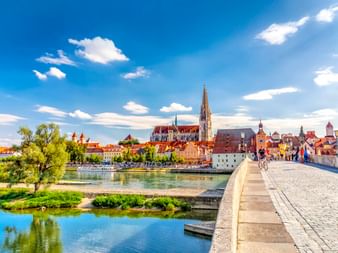 Steinerne Brücke über die Donau zur Regensburger Altstadt mit Domturm, bunten historischen Gebäuden und blauem Himmel.