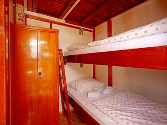 Kompakte Kabine auf der MS Orkan mit Holz-Etagenbetten, weißer Bettwäsche, einem Holzschrank und rot gestrichener Holzdecke und Wänden.