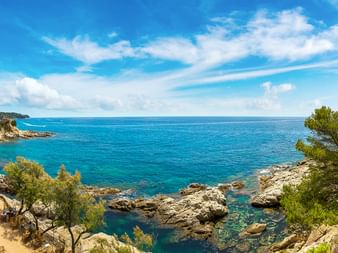 Panoramablick auf die Costa Brava Küste bei Lloret de Mar mit türkisfarbenem Mittelmeer, felsigen Klippen und Pinien unter blauem Himmel.