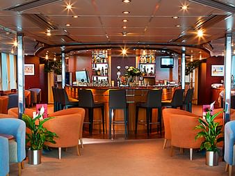 Geräumiger Salon an Bord der MS SE-Manon mit blauen und orangefarbenen Sesseln, zentraler Bar mit Hockern, großen Fenstern und warmer Beleuchtung.