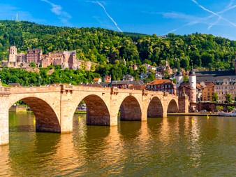 Historische Steinbrücke mit mehreren Bögen über den Neckar in Heidelberg, mit Schlossruine auf bewaldetem Hang und Kirchturm.