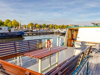 Sonniges Deck der MS Flora mit Holzbänken, weißem Geländer und Rettungsring. Marina mit Booten und Bäumen im Hintergrund unter blauem Himmel.