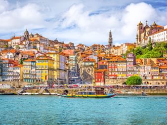 Bunte historische Gebäude von Porto erheben sich vom Douro-Flussufer. Traditionelle Boote auf blauem Wasser, Kirchtürme und orangefarbene Dächer unter bewölktem Himmel.