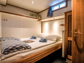 Gemütliche Doppelkabine auf der MS Flora mit gemachtem Bett, gestreifter Decke, Handtüchern, Holzschrank und Fenster mit Vorhang.
