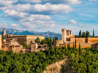 Panoramablick auf die Alhambra-Festung mit goldenen Mauern und Türmen, umgeben von grünen Bäumen, mit der Sierra Nevada im Hintergrund.
