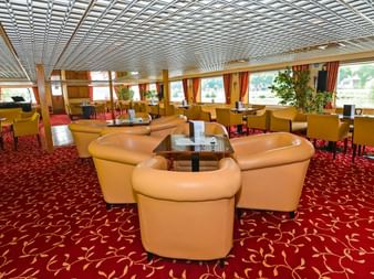 Geräumiger Salon an Bord der MS Normandie mit beigen Ledersesseln, rotem gemustertem Teppich und großen Fenstern mit orangen Vorhängen.
