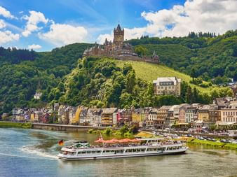 Reichsburg auf einem weinbewachsenen Hügel über der Stadt Cochem an der Mosel. Ein Ausflugsschiff fährt im Vordergrund auf dem Fluss.