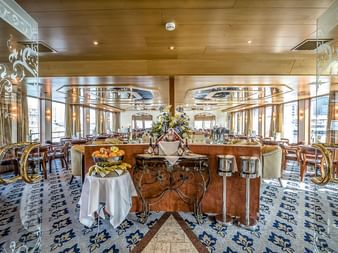Elegante Panoramabar auf der MS Swiss Crown mit Holzdecke, großen Fenstern, gemustertem Teppich und Buffettisch mit frischem Obst und Blumen.