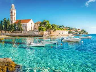 Franziskanerkloster mit Glockenturm an der Uferpromenade von Hvar. Kristallklares türkisfarbenes Wasser mit kleinen Booten im Hafen.
