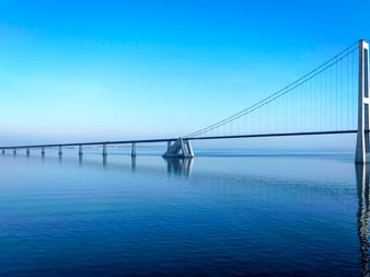 Große-Belt-Brücke über ruhigem blauen Wasser unter klarem Himmel. Die Hängebrücke mit weißen Türmen verbindet sich mit einer niedrigen Brücke.