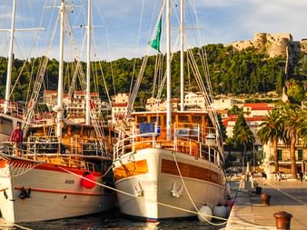 Vier hölzerne Kreuzfahrtschiffe am palmengesäumten Ufer in Kroatien. Historische Festung auf Hügel hinter Küstenstadt sichtbar.