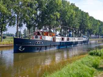 MS Magnifique II, ein blau-weißes Flusskreuzfahrtschiff mit Ankeremblem, fährt auf einem ruhigen Kanal gesäumt von hohen Bäumen und grünem Gras.