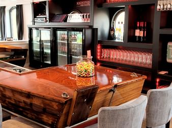 Moderne Bar an Bord der MS Magnifique III mit polierter Holztheke, gepolsterten Barhockern, Getränkekühlern und Weinregal. Große Fenster sorgen für Tageslicht.