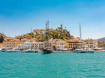 Hafen der Insel Poros mit weißen und gelben Gebäuden am türkisfarbenen Wasser. Glockenturm auf Hügel sichtbar. Segelboote und Yachten im Hafen.