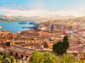 Aerial view of Portoferraio harbor with colorful buildings, terracotta roofs, ferries in the bay, and mountains in the background at sunset.