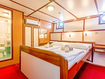 Schiffskabine auf der Amore mit Doppelbett, Holzverkleidung, rotem Teppich, Klimaanlage und eigenem Bad mit grünen Fliesen durch Tür sichtbar.