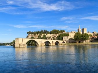 Historische Pont d'Avignon Brücke mit Steinbögen über die Rhône. Mittelalterlicher Papstpalast und Befestigungen auf dem Hügel unter blauem Himmel.