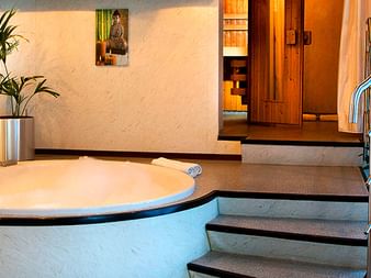 Wellnessbereich auf der MS SE-Manon mit rundem Whirlpool, Heimtrainer, grünen Pflanzen und Holzsauna im Hintergrund.