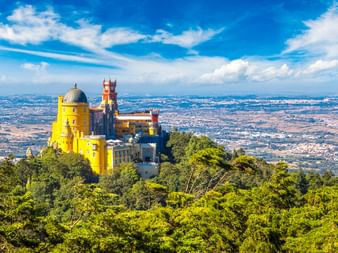 Palacio Nacional da Pena in Sintra mit leuchtend gelben und roten Mauern, umgeben von üppigem grünen Wald mit Panoramablick ins Tal unter blauem Himmel.