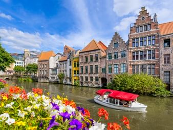 Ausflugsboot mit rotem Dach auf Kanal in Genter Altstadt, gesäumt von bunten mittelalterlichen Treppengiebelhäusern. Bunte Blumen im Vordergrund.