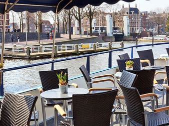 Sonnendeck der MS Fleur mit schwarzen Korbstühlen und Tischen mit Blick auf einen holländischen Kanal. Historische Gebäude und Boote am Wasser.