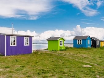 Reihe bunter Strandhäuser in Lila, Grün, Blau und anderen Farben auf grasbewachsener Küste von Ærø, Dänemark, mit Meer und bewölktem Himmel.