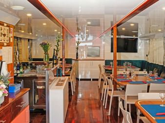 Innenraum des MS Azimut Salons mit Holzboden, gedeckten Tischen mit blauen und roten Tischsets, gepolsterten Bänken und Buffettheke mit Blumen.