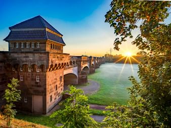 Historische Steinbrücke mit Turmgebäude über die Weser in Minden bei Sonnenaufgang, eingerahmt von grünen Bäumen mit goldenem Sonnenlicht.