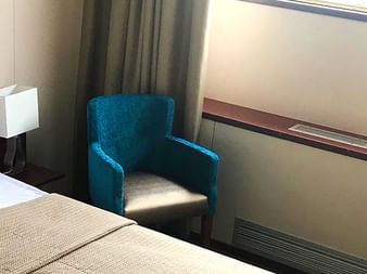 Kabine auf MS SE-Manon mit Doppelbett, weißer Bettwäsche, beigem Läufer, blauem Sessel, Nachttischlampe und großem Fenster mit Vorhängen.