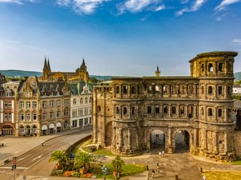 Porta Nigra, antikes römisches Stadttor in Trier mit verwitterten Sandsteinbögen. Historische Gebäude und Kirchtürme im Hintergrund.
