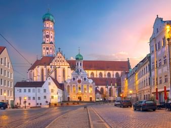Basilika St. Ulrich und Afra in Augsburg in der Dämmerung mit beleuchtetem Turm und Zwiebeldach. Kopfsteinpflasterplatz mit historischen Gebäuden.