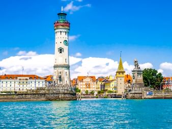 Weißer Leuchtturm mit grüner Spitze an der Lindauer Hafeneinfahrt am Bodensee. Bunte Gebäude und eine Löwenstatue am Ufer sichtbar.