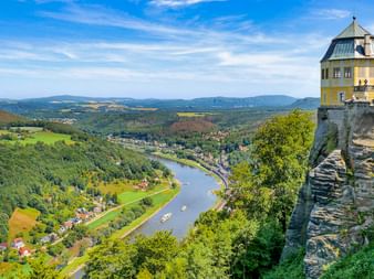 Gelber Barockturm auf Felsenklippe mit Blick auf das Elbtal mit grünen Wäldern, Feldern und fernen Bergen unter blauem Himmel.