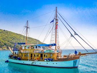 MS Panagiota, ein traditionelles Segelschiff mit blau-weißem Rumpf, vor Anker in türkisfarbenem Wasser bei Parga mit grünen Hügeln im Hintergrund.