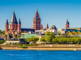 Blick auf die Mainzer Skyline vom Main mit dem roten Sandsteindom mit mehreren Türmen und modernen Gebäuden entlang der Uferpromenade.