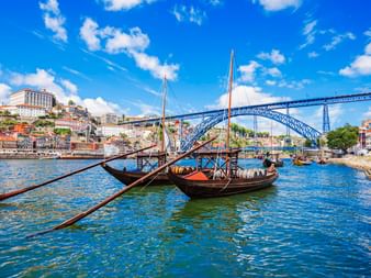 Traditionelle Rabelo-Boote mit Holzfässern auf dem Douro in Porto, mit der ikonischen Dom-Luís-I-Brücke und bunten Häusern am Hang.