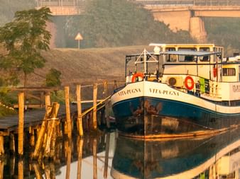 MS Vita Pugna, ein weiß-blaues Flusskreuzfahrtschiff, liegt an einem ruhigen Flussufer mit Holzpfählen vertäut. Radfahrer versammeln sich im Morgenlicht am Ufer.