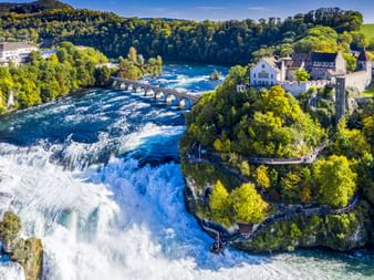 Luftaufnahme des Rheinfalls mit weißem Wasser, das über Felsen stürzt. Ein historisches Schloss und eine Brücke sind am rechten Ufer zu sehen, umgeben von grünen Wäldern.