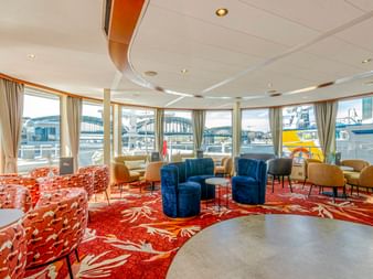 Geräumige Lounge auf der MS Lisabelle mit rot gemustertem Teppich, blauen und orangen Sesseln und großen Fenstern mit Hafenblick.