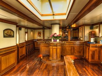 Eleganter Salon an Bord der MS Thalassa mit polierten Holzverkleidungen, zentralem Empfangstresen mit Blumen, Oberlichtern und gerahmten Kunstwerken.