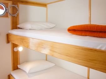 Kompakte Schiffskabine auf MS Planka mit Etagenbetten aus Holz, weißer Bettwäsche, orangen Kissen, Bullaugen und weißer Decke.