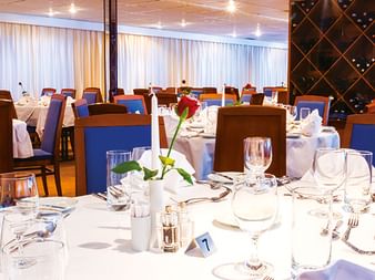 Elegantes Restaurant auf der MS SE-Manon mit weißen Tischdecken, blauen Stühlen und Weinregal. Tische gedeckt mit Gläsern und Blumen.