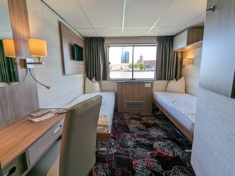 Kompakte Doppelkabine auf der MS Normandie mit zwei Einzelbetten, Holzverkleidung, Schreibtisch, Stuhl, Wandfernseher und Fenster mit Stadtblick.
