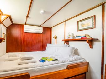 Kompakte Kabine auf der MS Caprice mit zwei Einzelbetten, weißer Bettwäsche, Holzverkleidung, Klimaanlage, Regal mit Büchern und gerahmten Bildern.