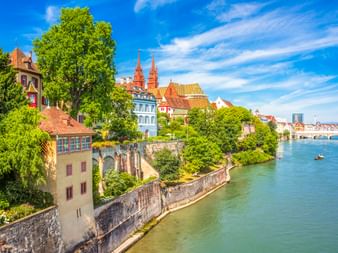 Basler Rheinufer mit bunten historischen Gebäuden, Kirchtürmen und Steinmauer entlang des Wassers. Üppige grüne Bäume säumen das Ufer unter blauem Himmel.